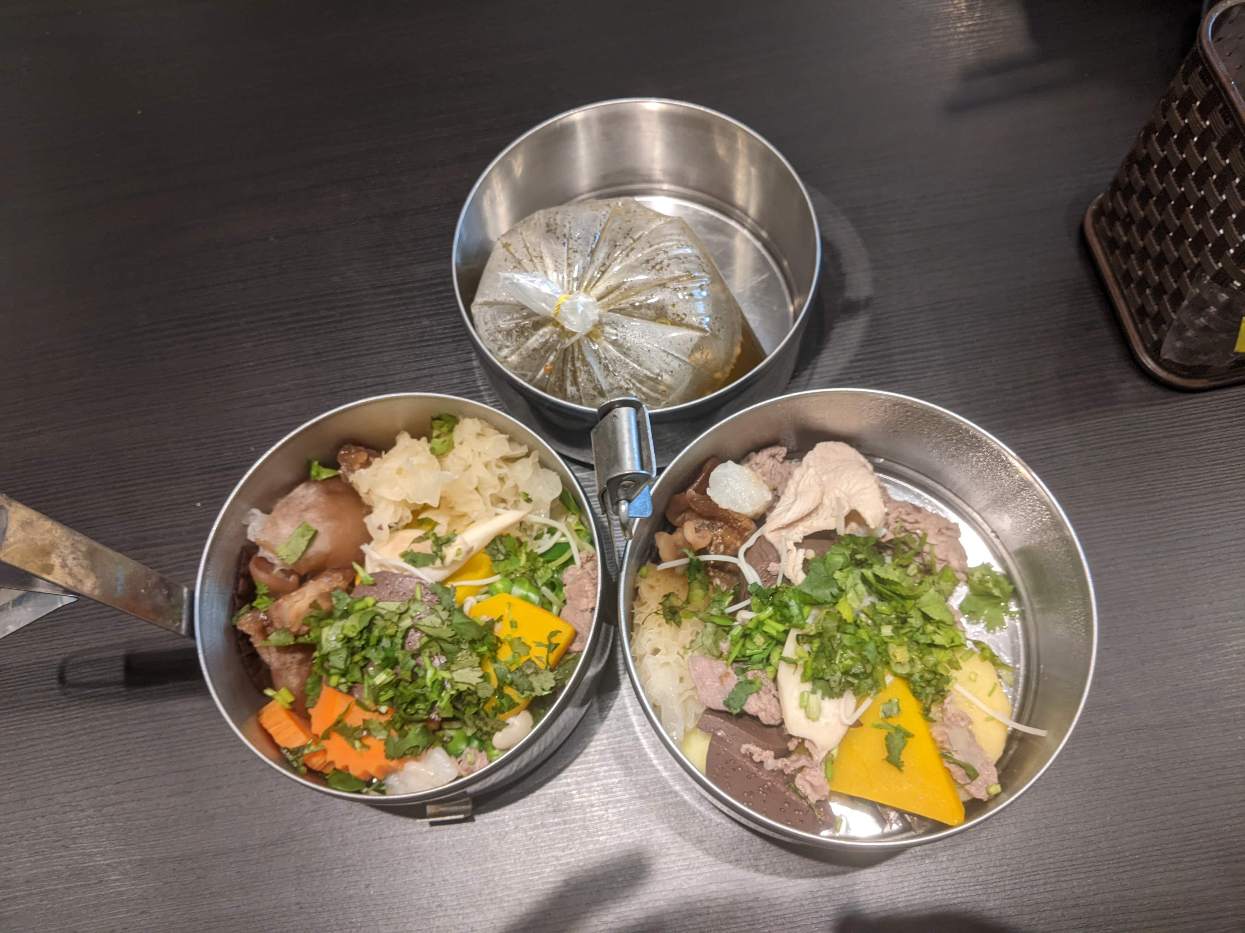 Two stainless steel food containers with boiled malatang ingredients and a separate soup bag in a plastic bag at Tak Tak Mala Chiang Mai